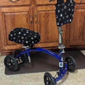 May include: A blue knee scooter featuring a black padded knee rest and a matching black storage pouch, both decorated with a white paw print pattern. The mobility device has four black wheels, a vertical handlebar stem, and a hand-operated brake system.