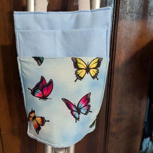 May include: A light blue fabric storage pouch with two top handles, featuring a front pocket printed with a pattern of colorful butterflies in shades of yellow, pink, blue, and orange against a soft, light blue sky-like background.