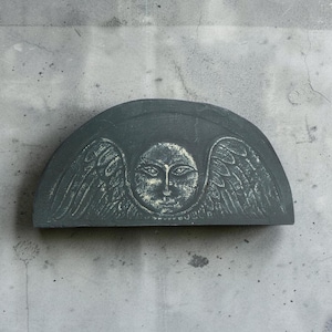 May include: A semi-circular dark gray stone plaque featuring a relief carving of a central face surrounded by stylized wings. The surface of the carving has a weathered, textured appearance with lighter gray highlights accentuating the details.