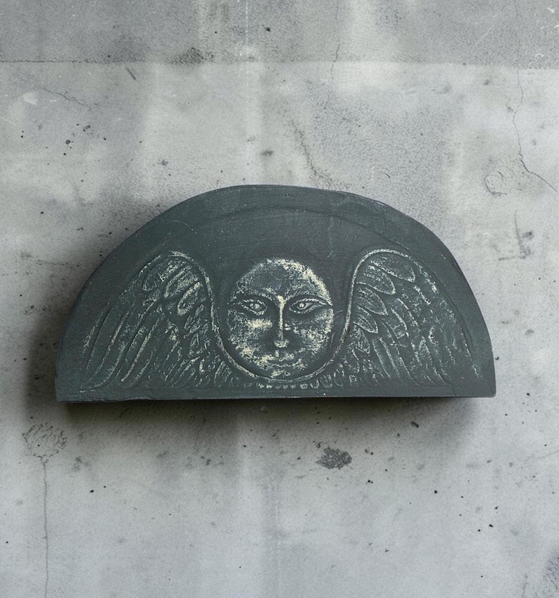 May include: A semi-circular dark gray stone plaque featuring a relief carving of a central face surrounded by stylized wings. The surface of the carving has a weathered, textured appearance with lighter gray highlights accentuating the details.
