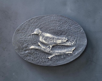 May include: An oval-shaped metal belt buckle featuring a raised relief design of a bird perched on a branch. The background has a textured, dark gray finish, while the bird and branch are highlighted in a polished, metallic silver tone.