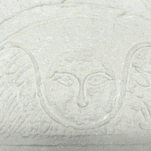 May include: A light beige stone relief carving featuring a central human face surrounded by stylized wings. The design is etched into a textured, matte surface with a semi-circular arch framing the figure, creating a subtle, monochromatic appearance.