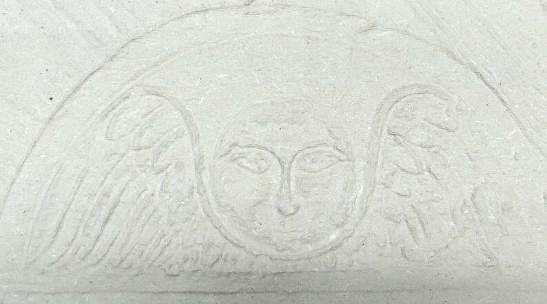 May include: A light beige stone relief carving featuring a central human face surrounded by stylized wings. The design is etched into a textured, matte surface with a semi-circular arch framing the figure, creating a subtle, monochromatic appearance.