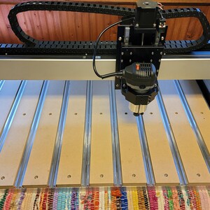 May include: A Shapeoko 4 CNC machine featuring a silver aluminum frame, a black spindle motor assembly, and a black cable management track. The work bed consists of parallel light brown MDF slats separated by silver metal channels with visible screws.