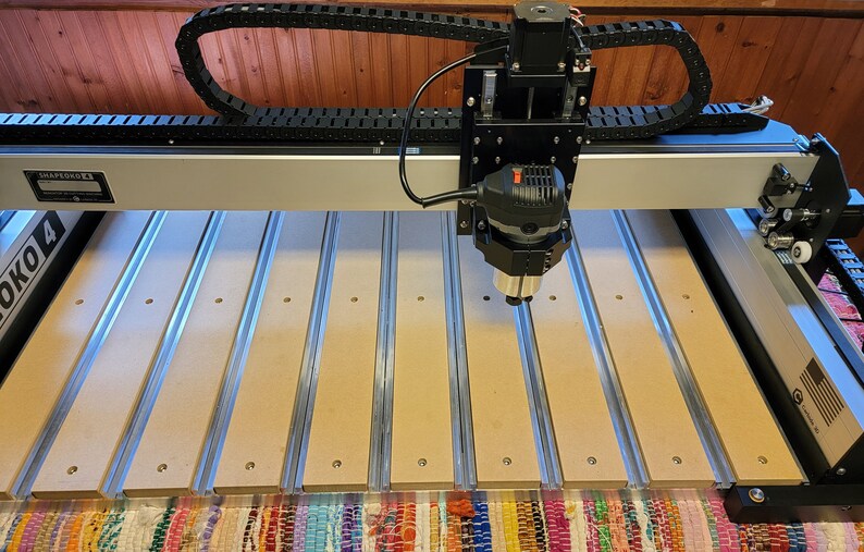 May include: A Shapeoko 4 CNC machine featuring a silver aluminum frame, a black spindle motor assembly, and a black cable management track. The work bed consists of parallel light brown MDF slats separated by silver metal channels with visible screws.