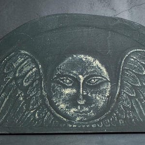May include: A semi-circular dark gray stone plaque featuring a carved relief of a central face surrounded by stylized wings. The engraved details are highlighted in a lighter, weathered white tone, creating a contrast against the dark surface.