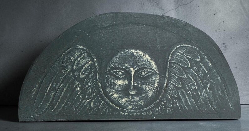 May include: A semi-circular dark gray stone plaque featuring a carved relief of a central face surrounded by stylized wings. The engraved details are highlighted in a lighter, weathered white tone, creating a contrast against the dark surface.