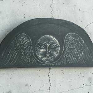 May include: A semi-circular dark gray stone plaque featuring a carved relief of a central face surrounded by stylized wings. The surface displays a weathered texture with lighter gray highlights accentuating the facial features and wing details.