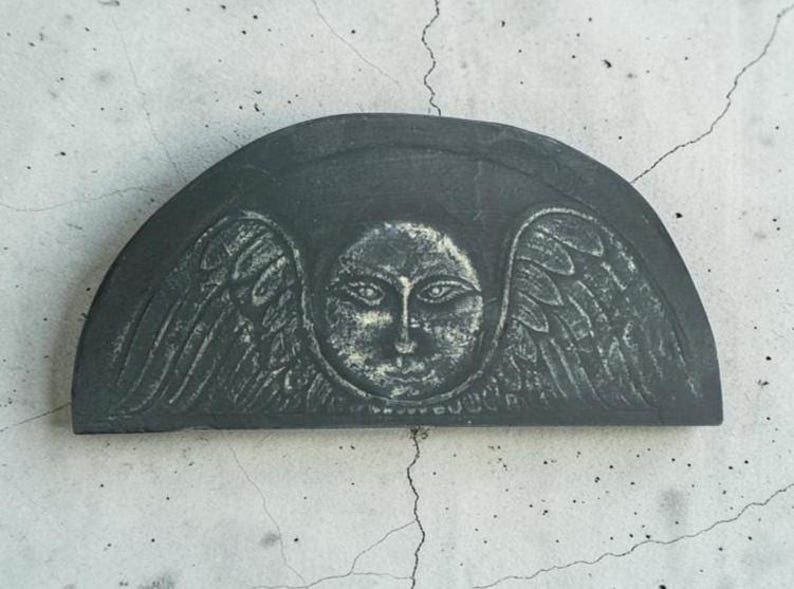 May include: A semi-circular dark gray stone plaque featuring a carved relief of a central face surrounded by stylized wings. The surface displays a weathered texture with lighter gray highlights accentuating the facial features and wing details.
