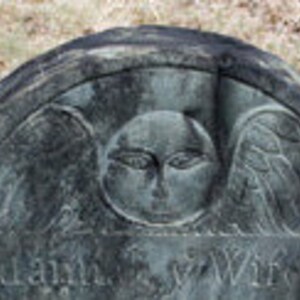May include: A weathered dark gray stone headstone featuring a carved winged cherub face at the top center. The arched top of the monument displays intricate relief carvings of wings extending from the central face, with partially visible text below.