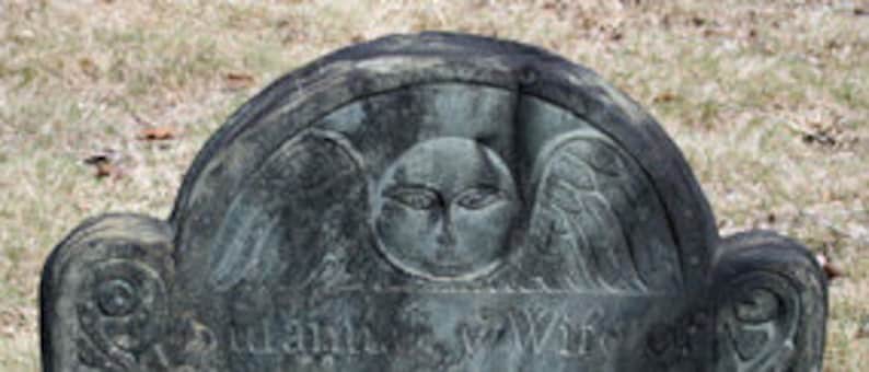 May include: A weathered dark gray stone headstone featuring a carved winged cherub face at the top center. The arched top of the monument displays intricate relief carvings of wings extending from the central face, with partially visible text below.