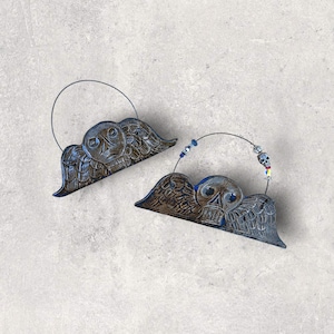 May include: Two decorative metal angel wing ornaments with skull faces. The wings are a mix of blue and brown, with a wire loop for hanging. One has a beaded accent with a skull charm.