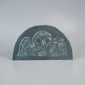 May include: A semi-circular dark gray plaque featuring a relief carving of a winged skull. The design includes a central skull with hollow eye sockets and a detailed set of teeth, flanked by stylized wings on both sides, all in a matte finish.