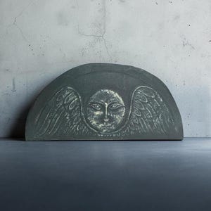 May include: A semi-circular dark gray stone plaque featuring a central carved face with detailed eyes and nose, flanked by symmetrical feathered wings. The surface has a weathered texture with lighter highlights accentuating the carved relief.