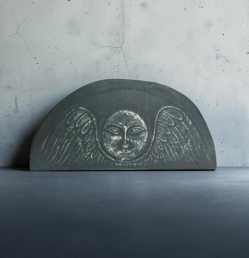 May include: A semi-circular dark gray stone plaque featuring a central carved face with detailed eyes and nose, flanked by symmetrical feathered wings. The surface has a weathered texture with lighter highlights accentuating the carved relief.