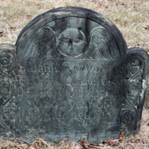 May include: A dark gray stone headstone with a rounded top featuring a carved winged face at the center. The surface displays weathered, partially legible engraved text and decorative scrollwork patterns along the left and right vertical edges.