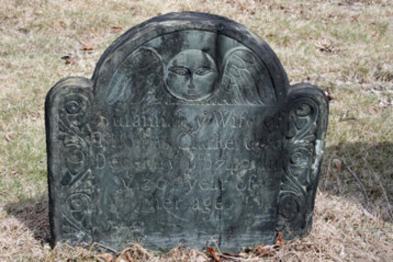May include: A dark gray stone headstone with a rounded top featuring a carved winged face at the center. The surface displays weathered, partially legible engraved text and decorative scrollwork patterns along the left and right vertical edges.