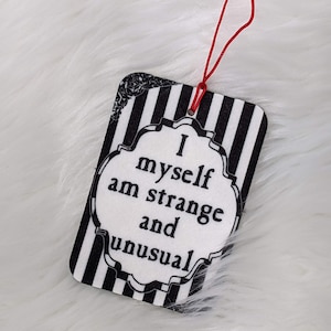 May include: Black and white car air freshener with the text "I myself am strange and unusual" in a white oval with a black border. The air freshener is hanging from a red string.