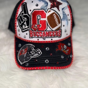 May include: A black and white trucker hat with a red and white design. The hat features the text "GO Buccaneers", a football, a star, and the number one hand symbol. Rhinestones and red trim add sparkle.