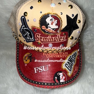 Op de afbeelding: Een op maat gemaakte baseballcap met een beige mesh achterkant en een rode klep. De cap heeft het Florida State Seminoles logo, een zwarte ster en de tekst "Game Day" en "FSU". Versierd met gouden studs en een gouden ketting.