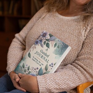 Könnte beinhalten: Ein rechteckiges geführtes Journal mit weißem Einband, das eine Aquarellillustration von blauen und violetten Blumen, grünen Blättern und Beeren zeigt. Der Text auf dem Einband lautet A Guided Journal for starting that next chapter of your life.