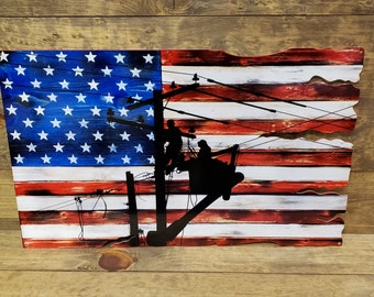 May include: Wooden American flag with a distressed design. The flag features the traditional blue field with white stars and red and white stripes. A black silhouette of utility workers on a power pole is overlaid on the flag.