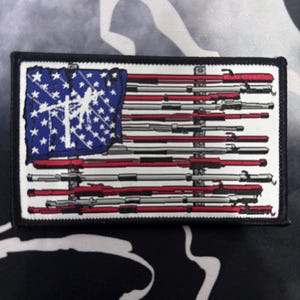 May include: A rectangular morale patch featuring a stylized American flag design. The blue canton contains white stars and a white rifle silhouette. The stripes are composed of alternating horizontal red and grey rifle barrels on a white background.