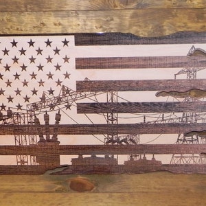 May include: A wooden American flag with a distressed, torn edge. The flag is stained in a dark brown color and features a silhouette of a construction site with cranes and buildings.