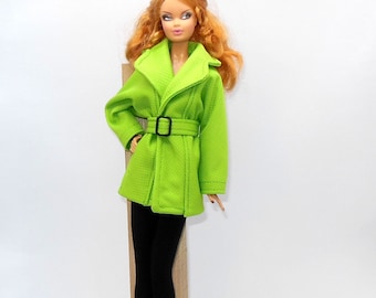 May include: A doll wearing a bright green coat with a black belt and black pants. The doll has red hair and sunglasses on top of its head. The coat has a textured pattern.