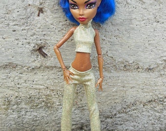 Può includere: Una bambola di moda con capelli blu e viola, che indossa un top e pantaloni dorati scintillanti. La bambola ha la pelle marrone e si trova in piedi contro un muro di cemento.