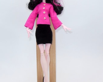Könnte beinhalten: Eine Modepuppe mit schwarzen Haaren und pinkfarbener und schwarzer Kleidung. Die Puppe trägt einen pinkfarbenen Pullover mit goldenen Knöpfen und einen schwarzen Rock. Die Puppe steht auf einer Holzplattform.