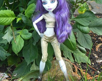 Può includere: Una bambola di moda con lunghi capelli viola, che indossa una canotta e pantaloni dorati. La bambola è in piedi in un giardino.