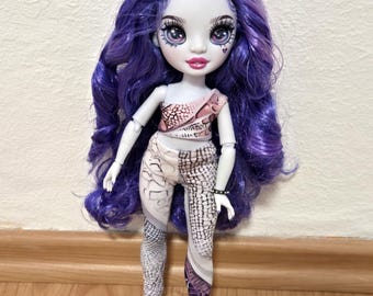 Può includere: Una bambola alla moda con lunghi capelli viola ondulati e lineamenti dettagliati, tra cui grandi occhi e labbra rosa. La bambola indossa un top bianco con un motivo rosa e nero e leggings abbinati. Un braccialetto nero adorna il polso della bambola.