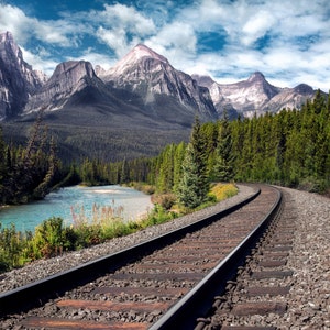 May include: A train track curves through a mountain valley with a river running through the center. The mountains are covered in trees and snow. The sky is blue with white clouds.