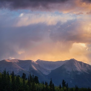 Könnte beinhalten: Eine Bergkette in dunkelblauem Farbton ist vor einem leuchtenden Sonnenuntergangs-Himmel silhouettiert. Der Himmel ist eine Mischung aus Rosa, Orange und Lila mit flauschigen weißen Wolken.