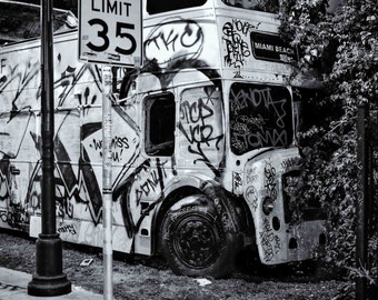 Puede incluir: Imagen en blanco y negro de un autobús de dos pisos cubierto de grafitis. El autobús tiene ventanas faltantes y está estacionado junto a una señal de límite de velocidad de 35. Las palabras "Miami Beach" son visibles.