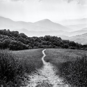 May include: A winding dirt path leads through a grassy field towards a distant mountain range. The sky is overcast with clouds, creating a dramatic black and white landscape.