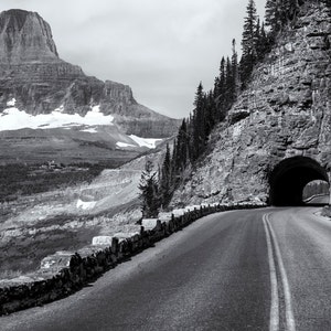 May include: A black and white photo of a winding road leading to a tunnel through a mountain. The road is lined with a stone wall and there is a snow-capped mountain in the background.