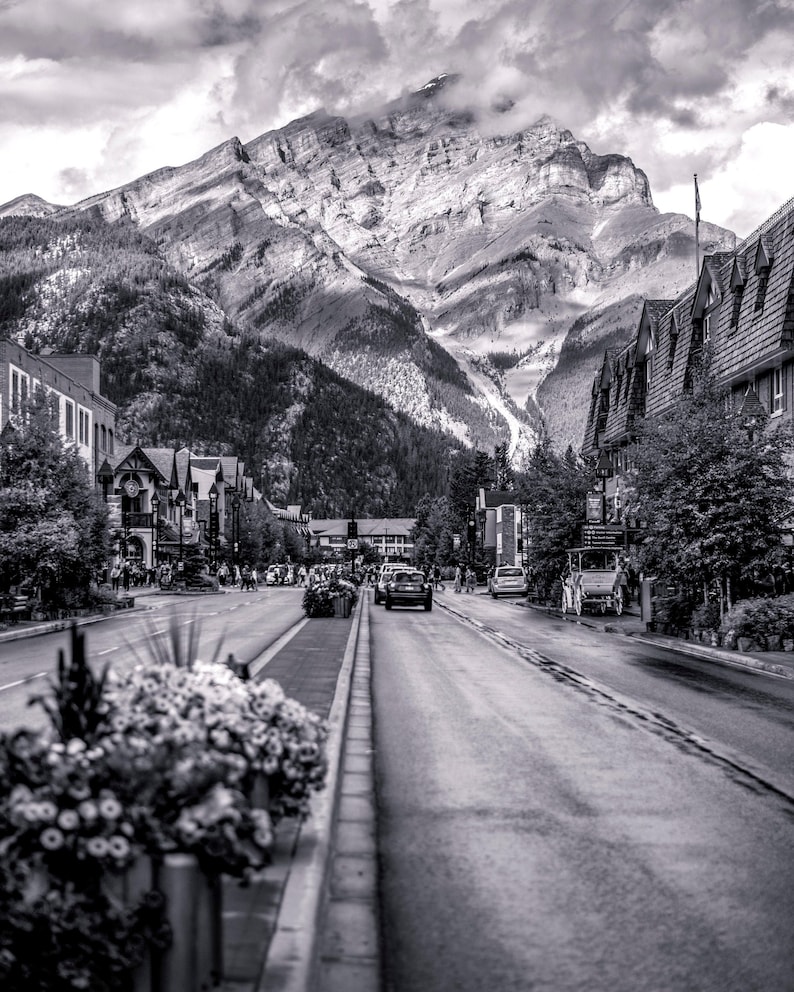 May include: A black and white photo of a street in a mountain town. The street is lined with buildings and trees. There are cars driving down the street and people walking on the sidewalk. In the background, a mountain range rises up in the distance.