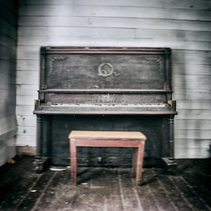 May include: A vintage upright piano with a wooden frame and ivory keys. The piano is in a room with white wood paneling and a wooden floor. A small wooden stool sits in front of the piano.
