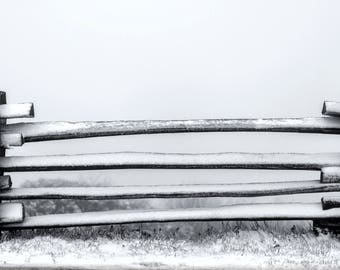 May include: A wooden fence with five horizontal rails covered in snow. The fence is made of rough-hewn logs and is set against a white, foggy background.