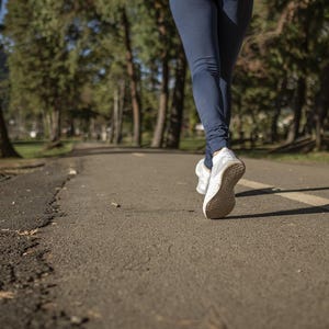 Könnte beinhalten: Nahaufnahme der Beine einer Person in marineblauen Leggings und weißen Turnschuhen, die auf einem asphaltierten Weg geht. Der Weg hat eine weiße Linie und ist von Bäumen und Gras umgeben.