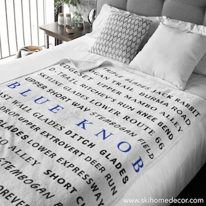 May include: White throw blanket with blue text that reads "Triple Glades Jack Rabbit", "Skyline Glades Lower Route 66", "D-Trail Ritchey's Run Knee Bend", "Edgeset Upper Mambo Alley", "Upper Short Way Stembogan Field", "Ogan Trail Burma Road", "Blue Knob", "St Wall Glades Ditch Glades", "Drop Upper Extrovert Deer Run", "Lades Lower Expressway", "O Alley Short C", "Tembogan R", "Reves". The blanket is on a bed with white sheets and gray pillows.