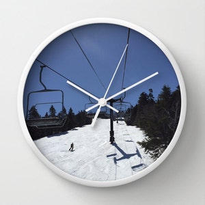 May include: A white clock with a blue sky and ski lift scene. The clock face shows a skier on a snowy slope with a ski lift in the background.