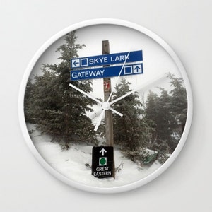 May include: A white clock featuring a snowy winter scene. The clock face shows a wooden pole with blue and white signs that read "Skye Lark" and "Gateway". A smaller sign at the bottom reads "Great Eastern".