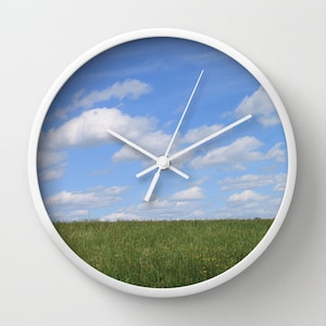 May include: White clock with a blue sky and white clouds image on the face. The clock hands are pointing to 3 o'clock. The bottom of the clock face shows a green grassy field.