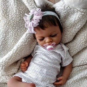 May include: A sleeping reborn baby doll with brown eyes, wearing a white and pink polka dot onesie and a pink and white headband with a flower. The doll is holding a pink pacifier.