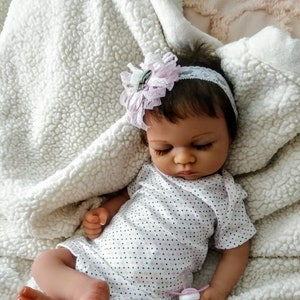 May include: A sleeping baby doll with brown hair wearing a white and black polka dot onesie and a pink flower headband. The doll is lying on a white fluffy blanket.