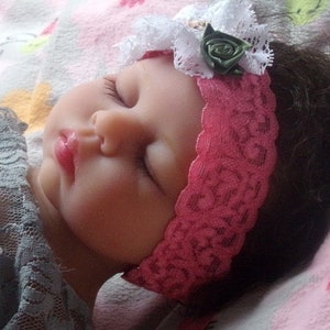May include: A sleeping baby doll with brown hair wearing a pink lace headband with a white lace flower and a green fabric rose.