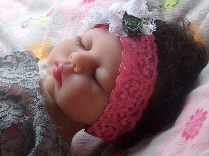 May include: A sleeping baby doll with brown hair wearing a pink lace headband with a white lace flower and a green fabric rose.